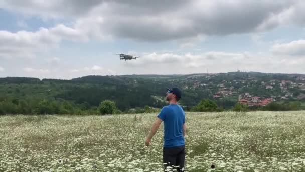 男性のドローンパイロットは 背景にかなりの町と美しい白い野の花の牧草地にドローンを着陸させます ブルガリアのNr Veliko Tarnovo — ストック動画