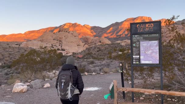 一位女徒步旅行者在日出时分从卡利科坦克小径出发 带着灿烂的山景 通过尾头标志和地图 美国内华达州红岩峡谷国家保护区 — 图库视频影像