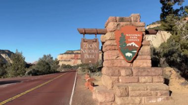 Güneşli bir günde Zion Ulusal Parkı 'nın doğu girişinin manzarası. Yeşiller yemyeşil ağaçlar ve çalılar, ABD 'nin Utah kentinin etrafını saran kayalıklarla parka giden kırmızı asfalt bir yol çiziyor.