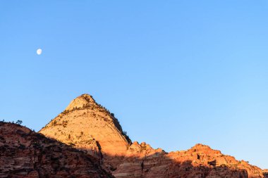 Parlayan kanyon duvarlarının manzarası kayalık tepelerde parlak güneş ışığı ve berrak mavi gökyüzüne karşı görünen bir ay. Serene manzarası doğanın güzelliğini yakalar - Zion Ulusal Parkı, Utah, ABD