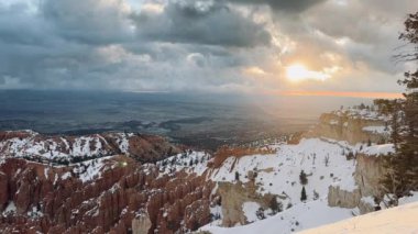 ABD 'nin Utah kentindeki Bryce Canyon Ulusal Parkı' nın kış manzarası. Güneş ışınları bulutların arasından karla kaplı kanyonu aydınlatıyor. Kırmızı kayalar ve çam ağaçları resimli bir manzara yaratır..