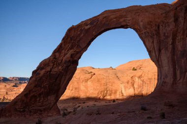 Moab, Utah 'taki Corona Arch' ta güzel bir gündoğumunun yakın çekimi, arka plandaki kırmızı kaya oluşumlarına sıcak bir ışık tutuyor. Doğal kemer mavi gökyüzüne çarpıcı bir siluet oluşturur.