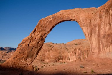 Gündoğumunda Moab, Utah 'taki Corona Kemeri' nin nefes kesici arka görüntüsü. Doğal kumtaşı kemeri, engebeli çöl manzarasıyla çevrili ılık bir parıltıyla berrak mavi bir gökyüzüne karşı duruyor.