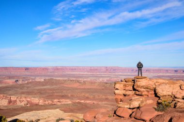 Yalnız bir adam, Utah 'taki dramatik Kanyonlar' ın geniş görüşlerini alan bir kayanın üzerinde duruyor. Açık mavi gökyüzü canlı kırmızı kaya oluşumlarını genişletir, macerayı sembolize eder - ABD