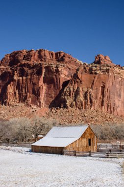 Utah 'taki Capitol Reef Ulusal Parkı' ndaki Fruita 'daki tarihi ahşap bir ahır. Karla kaplı manzara, parlak mavi gökyüzünün altındaki çarpıcı kızıl kaya kayalıklarıyla çelişiyor.