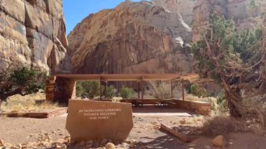 Capitol Resifi Ulusal Parkı patika başı. Bir piknik alanı kumtaşı kanyon duvarının ve patika işaretinin önünde duruyor. Tabela Petroglyphs Narrows, Pioneer Register ve Tanks 'ın Utah, ABD' deki izlerini gösteriyor.