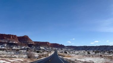 Güneşli bir kış gününde görüntülü araç kullanma. ABD 'nin Utah kentindeki Capitol Reef Ulusal Parkı' nda kar kaplı dağların ve kırmızı kaya oluşumlarının manzaralı 24 numaralı çevre yolunda bir araba..