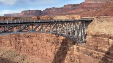 Bir Kaliforniya Akbabası, Arizona 'daki Mermer Kanyon' un nefes kesici güzelliğini sergileyerek Navajo Köprüsü 'nün altında zarafetle süzülür. Kızıl kaya kayalıklarının ve kemerli köprünün çarpıcı arkaplanı. 