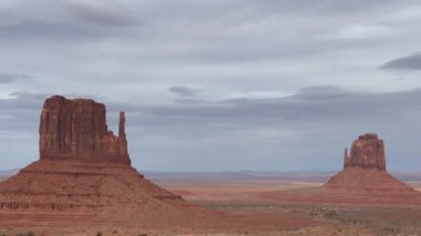 Arizona 'daki Anıt Vadisi' ndeki sembolik Mittens kırmızı kaya oluşumlarının geniş bir görüntüsü. Uçsuz bucaksız çöl manzarası bulutlu bir gökyüzünün altına yayılarak bir korku ve merak hissi yaratıyor.