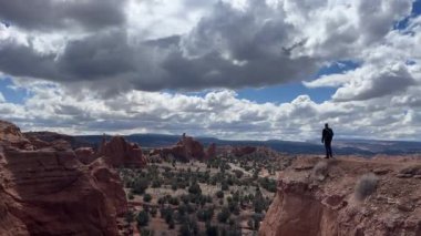 Çölde bir uçurumun kenarında duran bir adam, inanılmaz kaya oluşumlarına ve uçsuz bucaksız manzaraya bakıyor. Bulutlar mavi gökyüzüne çarpıyor. Kodachrome Havzası Eyalet Parkı, Utah, ABD