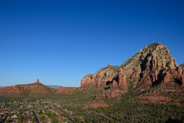 Sedona, Arizona 'nın nefes kesici görüntüsü, açık mavi gökyüzüne karşı duran ikonik kırmızı kaya oluşumlarını gözler önüne seriyor. Görüntü, dramatik çöl manzarasının arasına yerleşmiş sakin bir yerleşim alanı yakalıyor..