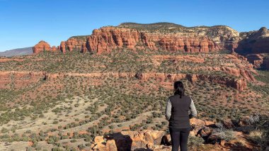 Bir kadın yürüyüşçü, Arizonas ikonik kızıl kaya dağları Sedona 'nın nefes kesici panoramik manzarasına hayranlıkla bakıyor. Engin çöl manzarası onun önünde uzanıyor.