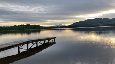 İskoçya 'daki Menteith Gölü üzerinde sakin bir günbatımı. Durgun suya uzanan ahşap iskele, renkli gökyüzünü ve uzak Ben Aan dağını yansıtıyor.