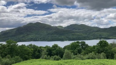 Katrine Gölü 'nün manzarası ve İskoçya' daki yemyeşil ağaçlar ve bitkilerle çevrili dağlar. Bulutlar mavi gökyüzünde sürükleniyor ve sakin atmosfere ekleniyor - Trossachs, İngiltere