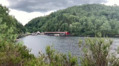 Lomond Gölü ve Trossachs Ulusal Parkı 'ndaki Katrine Gölü manzarası. Tekneli bir iskele ve bulutlu bir gökyüzünün altında orman yamacı sakin bir atmosfer yaratıyor.