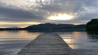 Ahşap iskele, resimli bir günbatımı sırasında İskoçya, Menteith Gölü 'ne uzanır. Sakin su, gökyüzünü ve uzak tepeleri yansıtarak İngiltere 'de sakin bir atmosfer yaratır.