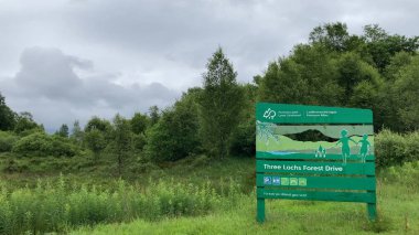 Aberfoyle, İskoçya, İngiltere - 07.10.2023: İskoçya 'daki Üç Lochs Forest Drive giriş tabelası ziyaretçileri ağırlıyor. Bereketli ağaçlar ve bulutlu gökyüzü huzurlu bir ortam oluşturur. Seyahat ve doğa içeriği için mükemmel..
