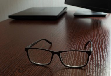 Stylish glasses on wooden office table close-up on blurred background of laptop and display