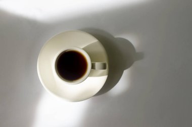 A cup of hot coffee in the morning sunlight  on white table. Hot drink in a mug for breakfast on background with window shadow. 