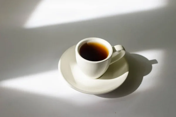 stock image A cup of hot coffee in the morning sunlight  on white table. Hot drink in a mug for breakfast on background with window shadow. 