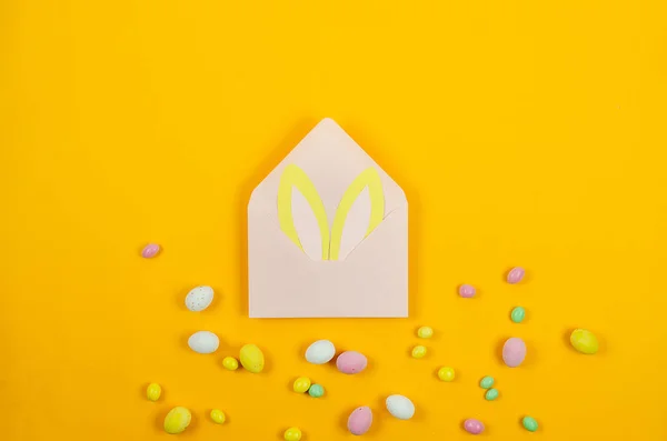 stock image Easter Colorful candy chocolate eggs and light pink envelope with blank card on yellow background. Easter holiday and tradition concept. Preparation for holiday. 