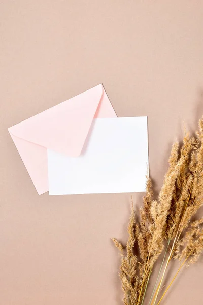 Pink envelope with empty white card for text, dry spikelets and decorative pumpkins on beige background. Blank invitation or greeting card mockup with copy space, autumn concept