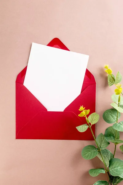 Red envelope with empty white card for text and eucalyptus branches on beige background. Blank invitation or greeting card mockup with copy space