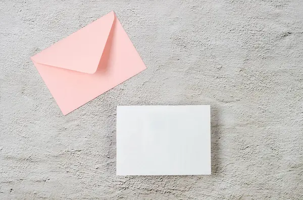 Pink envelope with empty white card for text on grey concrete background. Blank invitation or greeting card mockup with copy space