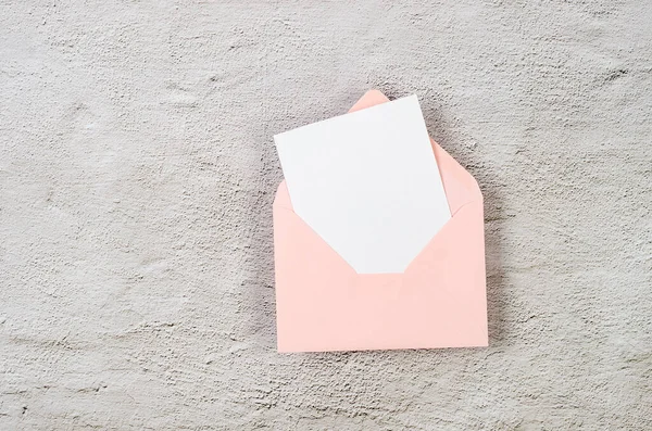 Pink envelope with empty white card for text on grey concrete background. Blank invitation or greeting card mockup with copy space