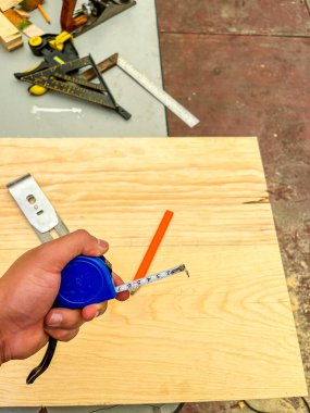 El, parmaklarının arasında mavi bir mezura tutuyordu ve arka planda masa ve iş aracının bir parçası vardı.