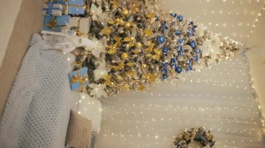 Dikey çekimde iki Noel ağacı altında hediyeler, kanepeler, duvarda çelenkler, odunlu şömine.