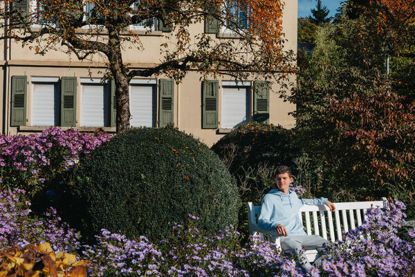 a teenager sits on a bench in the park drinks coffee from a thermo mug and looks into a phone. Portrait of handsome cheerful guy sitting on bench fresh air using device browsing media smm drinking