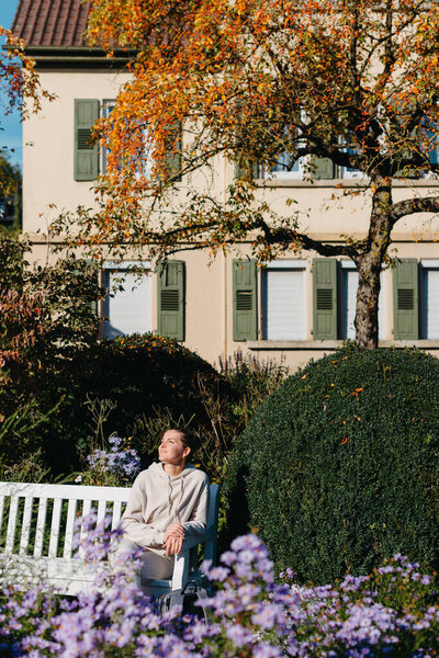 a girl sits on a bench in the park and enjoys the sun. Portrait young adult attractive woman enjoy sitting on bench and relaxing calm carefree rest in city park against green grass and trees on sunny