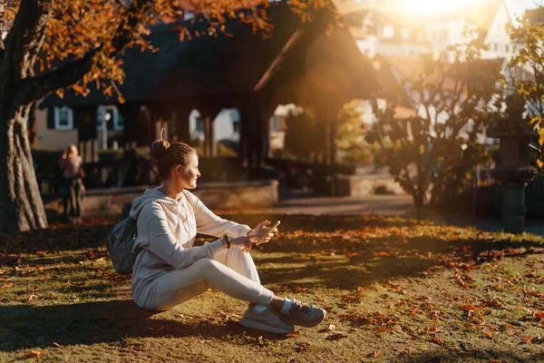 秋天的时候 年轻时髦的少女带着智能手机坐在公园里笑着 时尚的年轻女子在秋天的公园短信 重新触摸 充满活力的颜色 漂亮的金发少女穿休闲装 — 图库照片