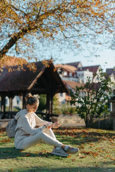 秋天的时候 年轻时髦的少女带着智能手机坐在公园里笑着 时尚的年轻女子在秋天的公园短信 重新触摸 充满活力的颜色 漂亮的金发少女穿休闲装 — 图库照片