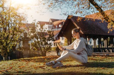 Sonbaharda parkta akıllı telefonlu genç ve şık bir kız gülümseyerek oturuyor. Sonbaharda parkta mesajlaşan modaya uygun bir genç kadın. Yenilenmiş, canlı renkler. Sıradan giyinen güzel sarışın genç kız.