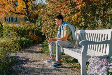 Parktaki bankta oturan bir genç termo bardaktan kahve içer ve telefona bakar. Yakışıklı, neşeli bir adamın portresi. Temiz hava almak için bankta oturuyor. Medya programında içki içiyor.