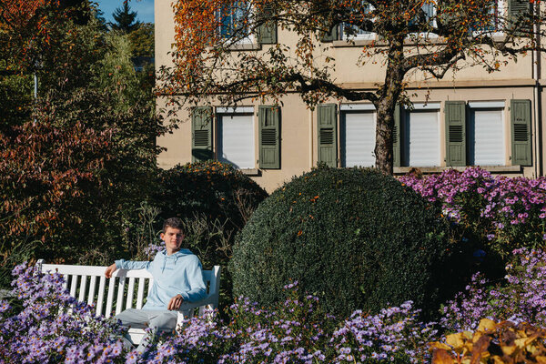 a teenager sits on a bench in the park drinks coffee from a thermo mug and looks into a phone. Portrait of handsome cheerful guy sitting on bench fresh air using device browsing media smm drinking