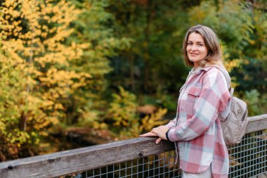 Sonbaharda gündelik giyinmiş genç bir kadının portresi, sonbahar parkının ve nehrin arka planında köprü üzerinde duruyor. Golden Fall 'da Park' ta yürüyen güzel bir kadın. Uzayı kopyala Gülümseyen kız