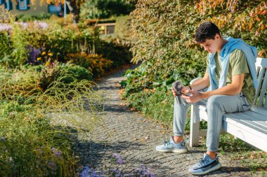 Parktaki bankta oturan bir genç termo bardaktan kahve içer ve telefona bakar. Yakışıklı, neşeli bir adamın portresi. Temiz hava almak için bankta oturuyor. Medya programında içki içiyor.