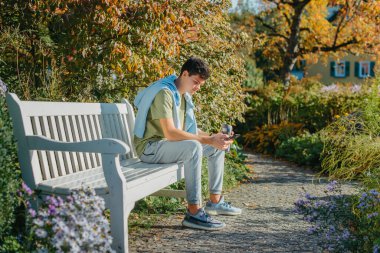 Parktaki bankta oturan bir genç termo bardaktan kahve içer ve telefona bakar. Yakışıklı, neşeli bir adamın portresi. Temiz hava almak için bankta oturuyor. Medya programında içki içiyor.