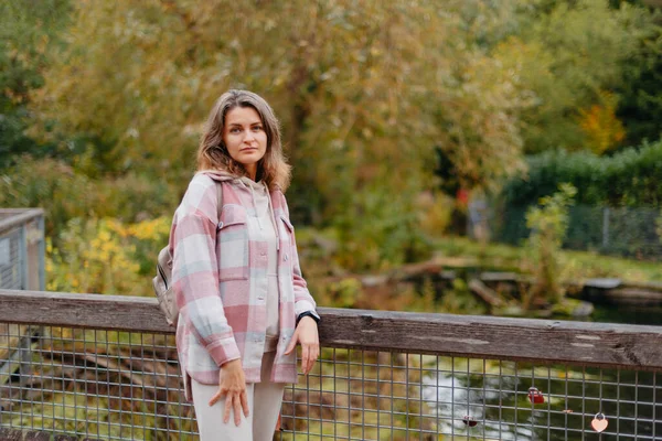 Portrait of cute young woman in casual wear in autumn, standing on bridge against background of an autumn Park and river. Pretty female walking in Park in golden fall. Copy space. smiling girl in the
