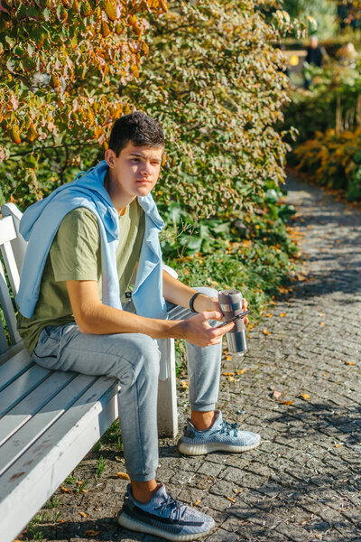a teenager sits on a bench in the park drinks coffee from a thermo mug and looks into a phone. Portrait of handsome cheerful guy sitting on bench fresh air using device browsing media smm drinking