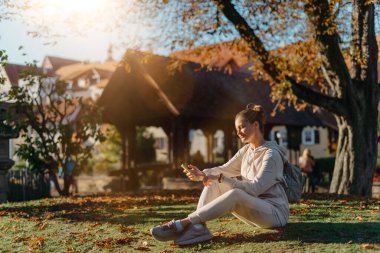 Sonbaharda parkta akıllı telefonlu genç ve şık bir kız gülümseyerek oturuyor. Sonbaharda parkta mesajlaşan modaya uygun bir genç kadın. Yenilenmiş, canlı renkler. Sıradan giyinen güzel sarışın genç kız.