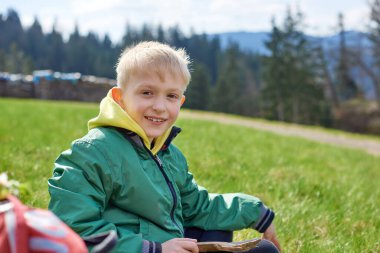 Eğlenceli genç yamaçta oturur. Çocuk dağlarda dinleniyor. Okul çocuğu çim tarlasında tepede oturuyor ve manzaranın tadını çıkarıyor. Doğada dinlenen ergen çocuk yürüyüşçüsünün arka görüntüsü. Etkin