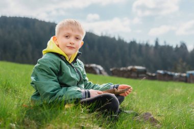 Eğlenceli genç yamaçta oturur. Çocuk dağlarda dinleniyor. Okul çocuğu çim tarlasında tepede oturuyor ve manzaranın tadını çıkarıyor. Doğada dinlenen ergen çocuk yürüyüşçüsünün arka görüntüsü. Etkin