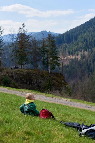 一个快乐的少年坐在山坡上 男孩在山上休息 小学生坐在草地上的小山上欣赏美丽的风景 少年徒步旅行者在大自然中休息的后视镜 积极活动 — 图库照片