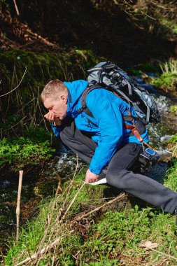 Yürüyüşçü dağda dere suyu içiyor. Yetişkin bir adam dağ deresinden su içiyor. Günlük giysiler ve güneş gözlüğü takan turist avuçlarından bir yudum nehir suyu içiyor.