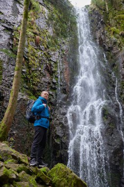 Almanya 'nın turistik merkezi - Schapbach yakınlarındaki Burgbach Şelalesi, Kara Orman, Baden-Wurttemberg, Almanya. Mavi ceketli bir adam taşın üzerinde duruyor ve düşen suyun akışına bakıyor.