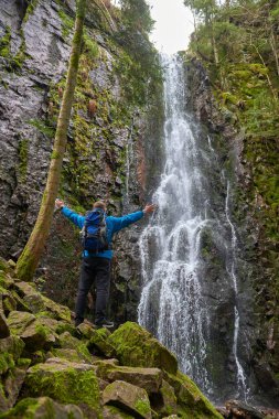 Almanya 'nın turistik merkezi - Schapbach yakınlarındaki Burgbach Şelalesi, Kara Orman, Baden-Wurttemberg, Almanya. Mavi ceketli bir adam taşın üzerinde duruyor ve düşen suyun akışına bakıyor.